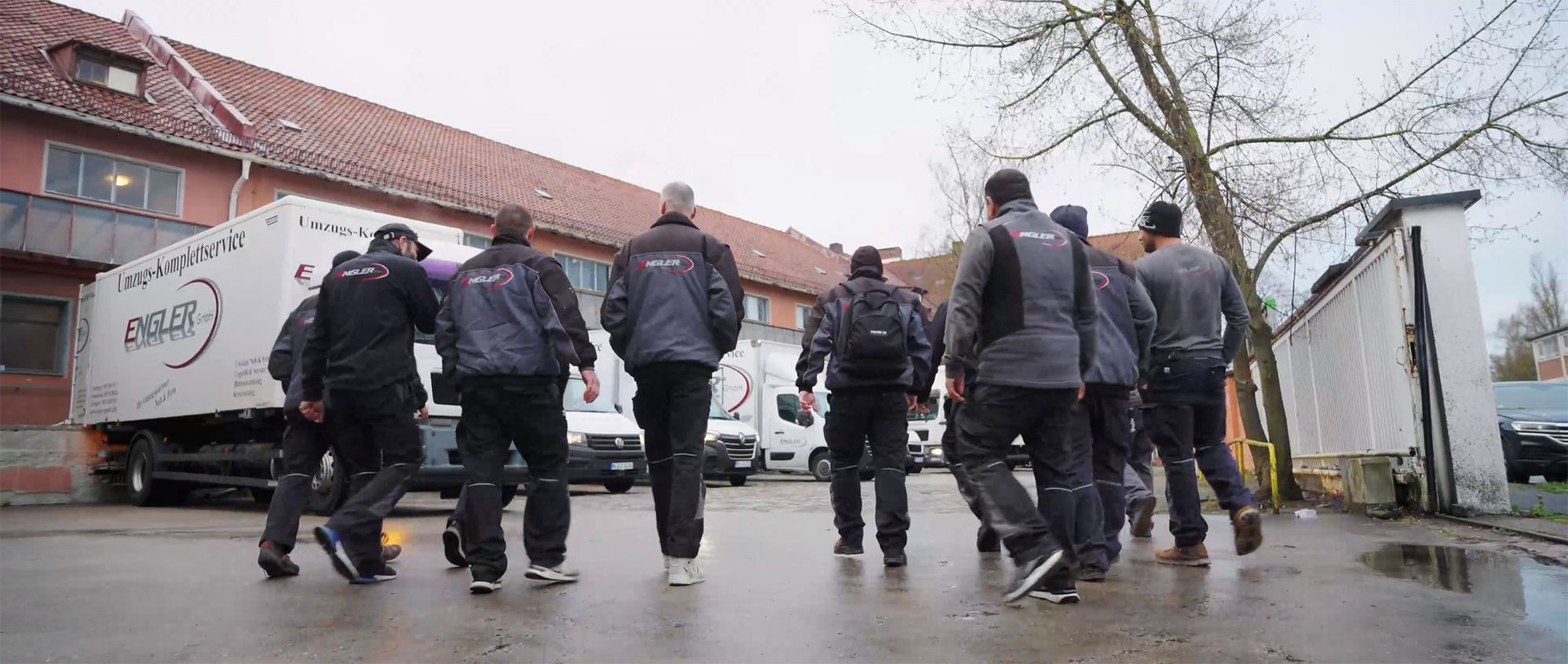 Sieben Personen in Arbeitskleidung mit dem Schriftzug Engler auf dem Rücken gehen auf mehrere Umzugsfahrzeuge zu, die vor einem Gebäude mit rotem Ziegeldach stehen.