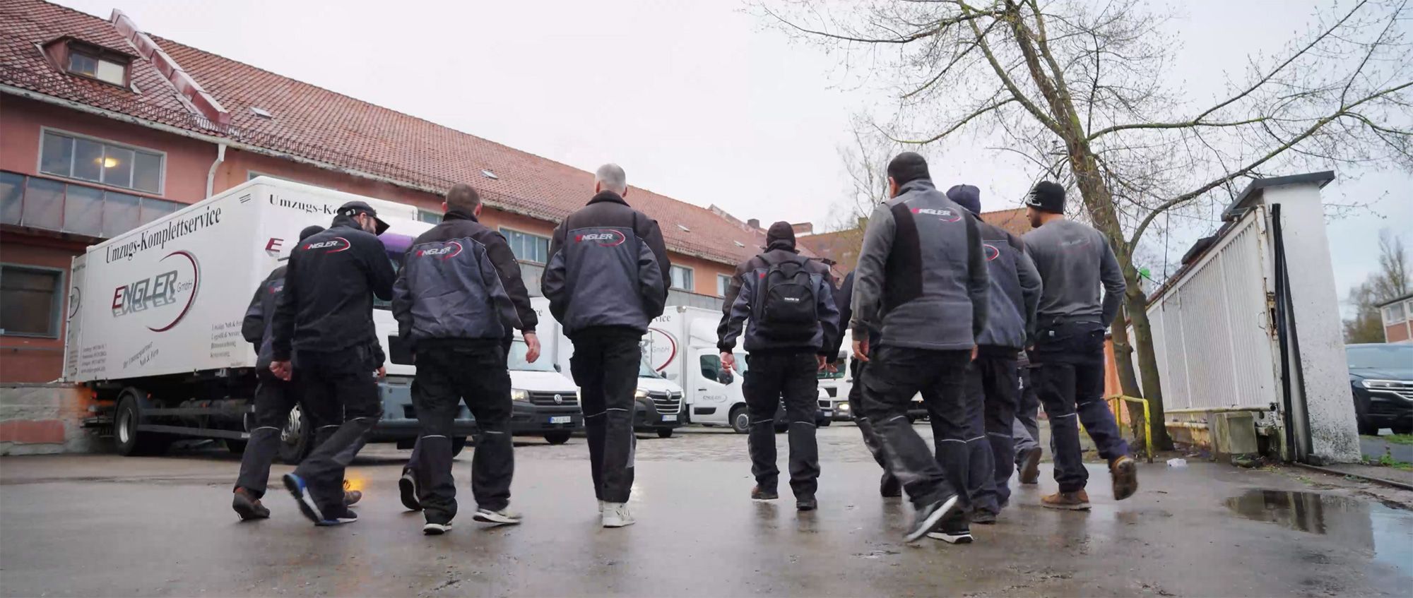 Sieben Personen in Arbeitskleidung mit dem Schriftzug Engler auf dem Rücken gehen auf mehrere Umzugsfahrzeuge zu, die vor einem Gebäude mit rotem Ziegeldach stehen.