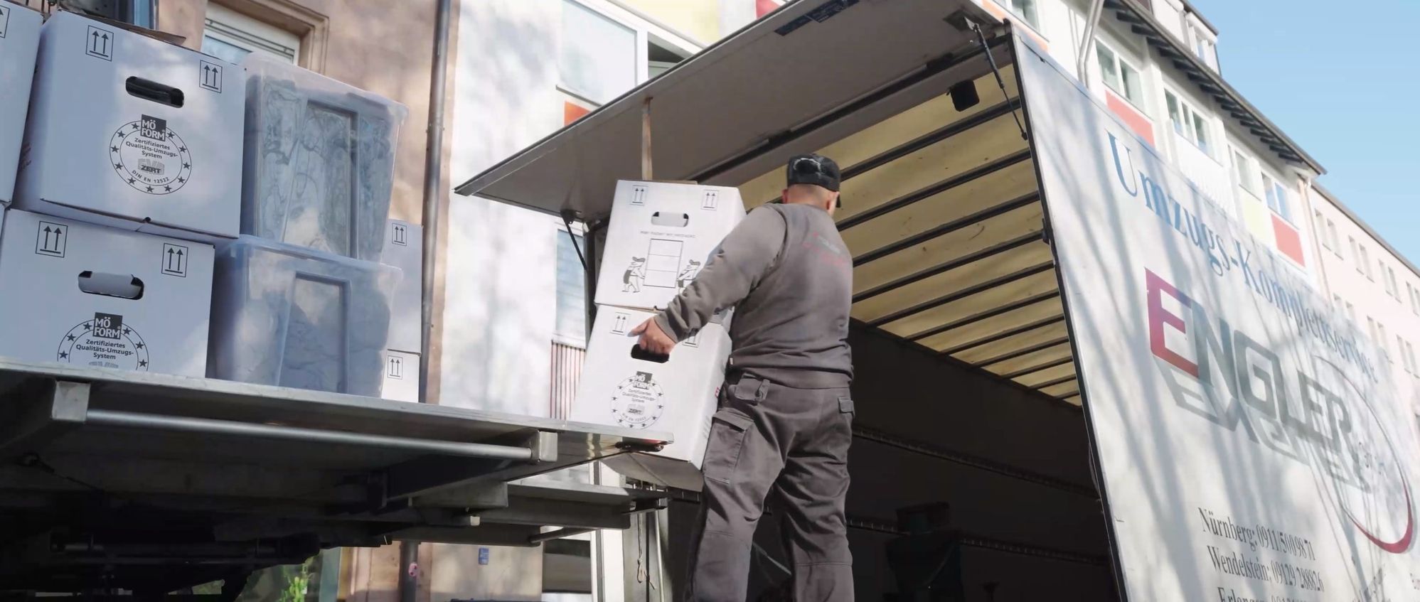 Mann hebt Karton mit Aufdruck vor geöffnetem Umzugs-Lkw der Engler GmbH in einem Wohngebiet