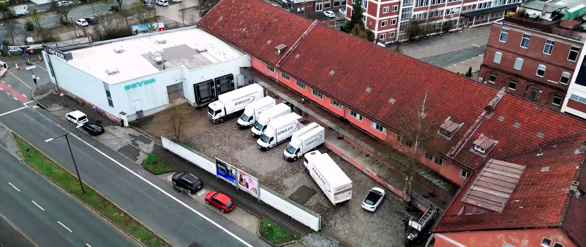 Luftaufnahme eines Firmengeländes mit mehreren weißen Umzugs-Lkw und einem roten sowie einem schwarzen Pkw vor Gebäuden mit roten Ziegeldächern