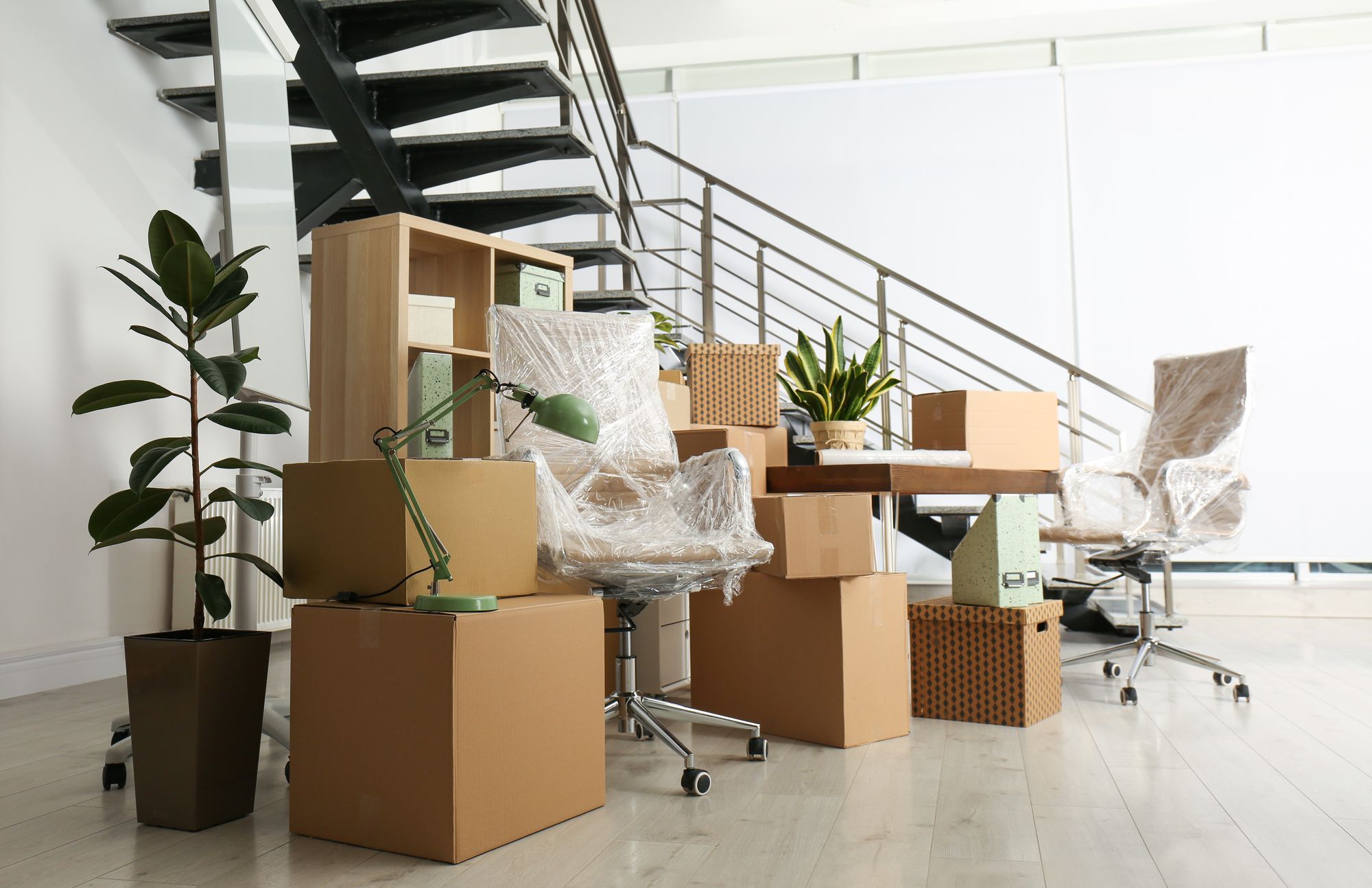 Büro mit verpackten Stühlen, Umzugskartons, Schreibtischlampe und Pflanzen vor einer Treppe