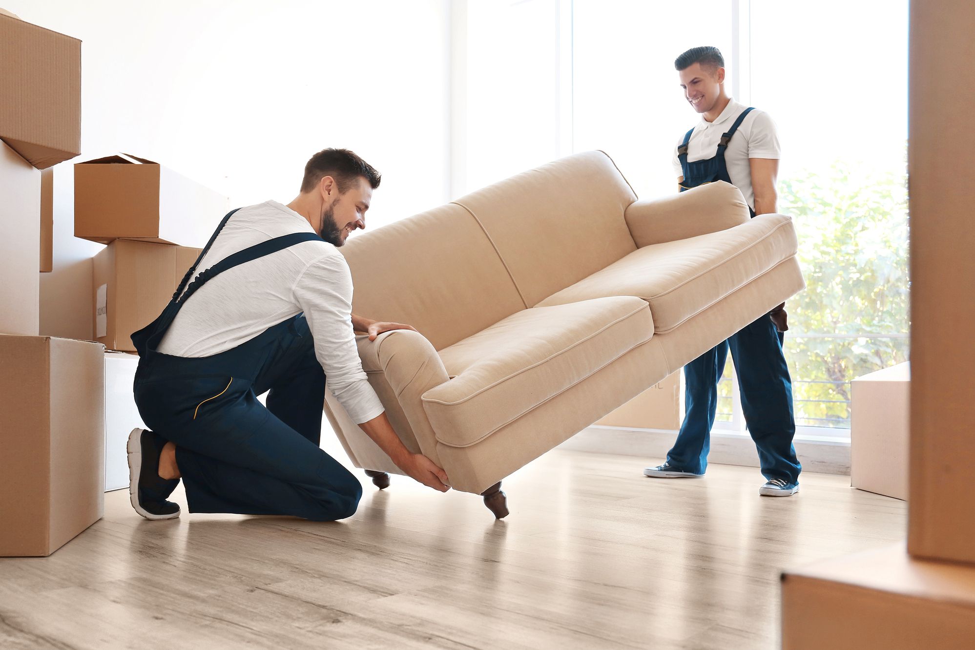 Zwei Männer in Arbeitskleidung heben gemeinsam ein Sofa in einem hellen Raum mit Umzugskartons